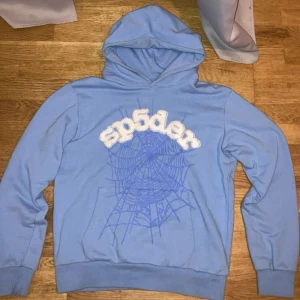 spider  - Säljer en ljusblå hoodie från Sp5der med stort vitt broderat logotyp-tryck och blått spindelnätsmönster på bröstet. Tröjan har huva och magficka, samt ribbade muddar vid ärmslut och nederkant. Perfekt för dig som gillar streetwear vibes. Möts bara upp!