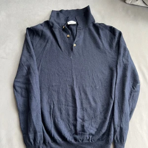 Mörkblå stickad tröja med knappar - Mörkblå stickad tröja från Massimo Dutti med hög krage och tre snygga knappar framtill. Tröjan har långa ärmar och ribbade muddar vid ärmslut och nederkant. Perfekt för dig som gillar en klassisk och stilren look.