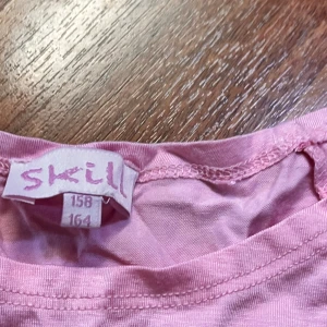Rosa kortklänning från Skill - Söt rosa kortklänning från Skill med långärmade ärmar och rund hals. Överdelen är i mjuk bomull och kjolen har ett skirt lager som ger en fluffig känsla. Perfekt för dig som gillar en feminin och lekfull stil.