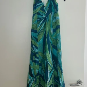 Grönblå långklänning med knyt - Snygg långklänning med tropiskt mönster i grönt, blått och turkost. Klänningen har v-ringad hals och knyt i nacken, vilket ger en cool halterneck-look. Materialet är lätt och luftigt, perfekt för varma dagar. Passformen är loose och klänningen är ärmlös.