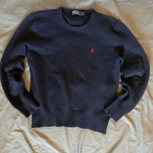 Mörkblå ulltröja från Polo Ralph Lauren - Mörkblå stickad tröja från Polo Ralph Lauren med klassisk röd broderad logga på bröstet. Tröjan har rund halsringning, ribbade muddar och är tillverkad i mjuk ull. Perfekt för lager-på-lager under kyliga dagar.
