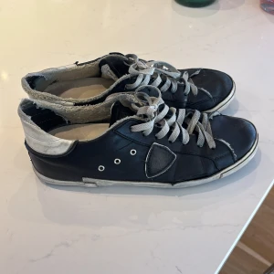 Svarta Philippe modell - Svarta sneakers med grå snörning och vit/grå häl. Skorna har rund tå, platt sula och flera metallöljetter på sidan. Ovandelen är i skinn och de har en cool, sliten look med kontrasterande detaljer på hälen och sidan. Snygga men använda hör av er vid funderingar. Har en del defekter så skriv för att se dem. Box följer med.
