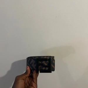 Brunt Fendi bälte med FF-logga - Säljer ett snyggt bälte från Fendi i brunt och svart med det klassiska FF-mönstret över hela bandet. Bältet har ett brett svart spänne i metall som verkligen sticker ut. Perfekt accessoar för att lyfta din outfit med en lyxig touch.