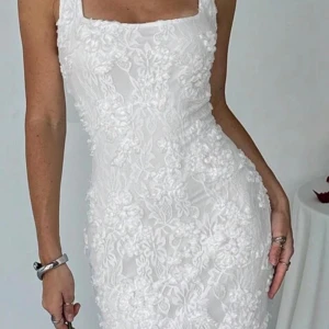 Vit blommig spetsklänning med smala axelband - Supersnygg vit klänning med smala axelband och figurnära passform. Klänningen har ett blommigt spetsmönster över hela och en fyrkantig urringning framtill. Perfekt för dig som vill ha en romantisk och elegant look med extra detaljer.