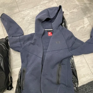 Mörkblå Nike hoodie med dragkedja - Säljer en mörkblå hoodie från Nike med hel dragkedja och huva. Tröjan har långa ärmar, två fickor med dragkedja och diskret Nike-logga på bröstet. Perfekt för chill dagar eller när du vill ha en sportig look.