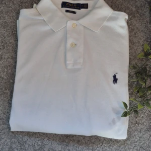 Vit pikétröja Polo Ralph Lauren M - Vit pikétröja från Polo Ralph Lauren i slim fit-modell. Klassisk krage, två knappar och den ikoniska broderade loggan i mörkblått på bröstet. Tillverkad i 100% bomull för en skön och luftig känsla.