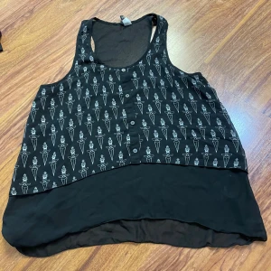 Svart linnetopp med mönster från Divided - Svart linnetopp från Divided med coolt vitt grafiskt mönster framtill och knappar längs bröstet. Toppen är ärmlös och har en längre, rundad nederkant i tunt, flowy material. Perfekt för dig som gillar edgy och unik stil.