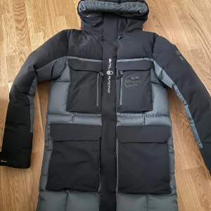 Sail Racing dunjacka svart/grå - Säljer en fet dunjacka från Sail Racing i svart och grått med stora fickor, huva och coola loggor på ärm och rygg. Jackan har dragkedjor, GORE-TEX Infinium-tyg och är perfekt för kalla vinterdagar. Snygg, lång modell med riktigt skön passform och stilren design.