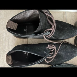 Svarta mockakängor Hansen & Jacob - Snygga svarta kängor från Hansen & Jacob i mocka med rund tå och snörning. Yttersulan är i läder och gummi för extra grepp. Perfekt för dig som gillar stilrena och klassiska skor med en modern touch.