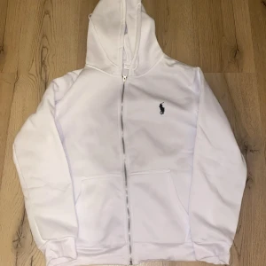 Vit hoodie från Polo Ralph Lauren - Vit hoodie från Polo Ralph Lauren med dragkedja och huva. Klassisk design med broderad marinblå logga på bröstet, fickor framtill och snörning i huvan. Mjuk och skön bomullskvalitet, perfekt för chill dagar.