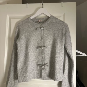 Grå kofta med rosett-detaljer från Nelly - Mysig ljusgrå kofta från Nelly med rund hals och långa ärmar. Framtill har den tre söta rosett-detaljer och knappar. Materialet är stickat och känns mjukt mot huden. Perfekt för att addera lite gullig stil till din outfit.