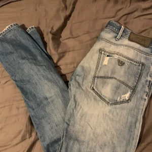 Ljusblå distressed jeans från Armani - Säljer ett par ljusblå jeans från Armani med snygga slitningar och patchade detaljer. Klassisk femficksmodell med logga på bakfickan och normal passform. Jeansen är tillverkade i bomull och har en modern, streetinspirerad look.