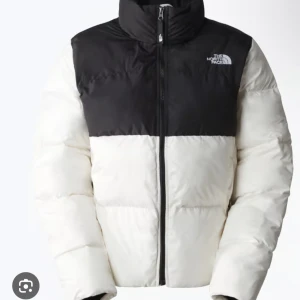 Svartvit pufferjacka från The North Face - Säljer en snygg pufferjacka från The North Face i svart och vitt. Jackan har hög krage, dragkedja framtill och logga på bröstet. Perfekt för kalla dagar med sin fluffiga och isolerande design. Passar dig som gillar streetstyle och vill hålla värmen.