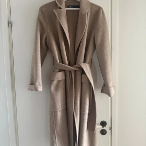 Beige kappa med bälte från Zara  - Stilren beige kappa från Zara i storlek XS. Lång modell med klassisk krage, stora fickor och knytskärp i midjan. Perfekt för dig som gillar en clean look och vill ha en jacka som är både snygg och bekväm. Har inte använt denna jackan mycket så materialet är som nytt✨