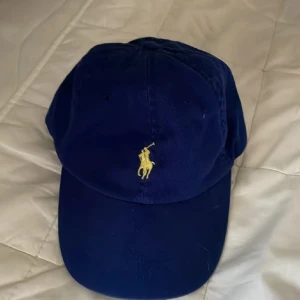 Blå keps från Polo Ralph Lauren - Snygg marinblå keps från Polo Ralph Lauren med klassisk gul broderad logga framtill och 'POLO' broderat bak. Justerbart spänne baktill för perfekt passform. Tillverkad i 100% bomull, perfekt för en clean och sportig look.