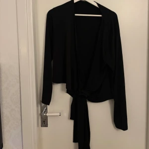 Svart omlottopp med knyt - Snygg svart omlottopp med långa ärmar och knyt framtill. Toppen har en croppad passform och är perfekt för dig som gillar stilrena plagg med lite edge. Materialet känns mjukt och följsamt, vilket gör den skön att bära. Enkel att matcha med både jeans och kjol.