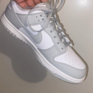 Nike Dunk Low vit och ljusgrå - Nike Dunk Low sneakers i fräscht vitt och ljusgrått skinn. Klassisk siluett med rund tå, perforerad tåbox och platt sula. Snygga grå skosnören och ikoniska Swoosh-loggan på sidan. Perfekta för dig som gillar stilrena och cleana sneakers.