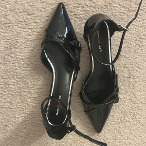 Svarta lackpumps från Bata Red Label - Snygga svarta pumps i lack från Bata Red Label med spetsig tå, låg klack och dubbla smala remmar med spännen över foten. Skorna har en elegant siluett och anklerem för extra support. Perfekta för dig som vill sticka ut med en klassisk men edgy look.