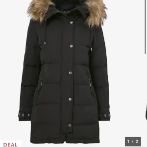 Svart lång dunjacka med fuskpäls - Snygg svart dunjacka från Hollies med lång passform och avtagbar huva med fuskpäls. Jackan har dragkedja och knappar framtill, två fickor med dragkedja och justerbara muddar vid ärmslut. Perfekt för kalla vinterdagar.