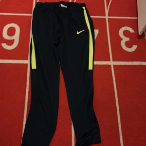 Nike Dri-Fit träningsbyxor M - Svarta Nike Dri-Fit träningsbyxor med gula ränder längs sidorna och snörning i midjan. Byxorna har en sportig look och är gjorda i ett lätt och funktionellt syntetmaterial som andas. Perfekta för gymmet eller löpning.