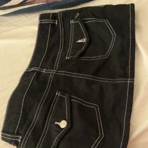 Svart minikjol med kontrastsömmar - Svart kortkjol med vita kontrastsömmar och stora fickor framtill. Kjolen har knapp och dragkedja i midjan samt bälteshällor. Snygg streetstil med markerade detaljer fickor.