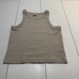 Beige ribbad linnetopp från Lindex - Säljer en beige ribbad linnetopp från Lindex i storlek 146/152. Toppen är ärmlös med klassisk rund halsringning och har en enkel, stilren design. Perfekt för varma dagar eller som lager under andra plagg.