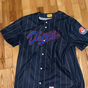 Randig basebolltröja från Dime - Säljer en mörkblå basebollinspirerad tröja från Dime med vita ränder, broderad logga på bröstet och patch på ärmen. Tröjan har knappar framtill och är kortärmad. Perfekt för dig som gillar streetwear och sportig stil.