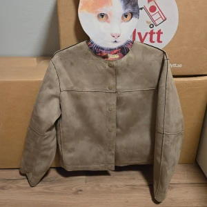 Beige mockajacka med tryckknappar - Snygg beige mockajacka med raka linjer och tryckknappar framtill. Jackan har en minimalistisk design, lång ärm och markerade sömmar som ger en clean look. Perfekt för dig som gillar stilren och enkel vibe.