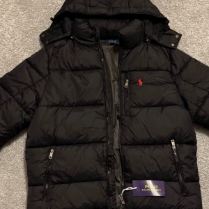 Polo Ralph Lauren puffer/dunjacka - Svart dunjacka från Polo Ralph Lauren med huva, dragkedjor i silver och klassisk röd logga på bröstet. Jackan är quiltad och har flera fickor med dragkedja. Perfekt för kalla dagar och riktigt snygg streetstil.