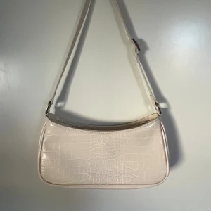 Beige axelväska i krokopräglat syntet - Snygg beige axelväska med krokopräglad yta och justerbar rem. Väskan har silvriga metalldetaljer och dragkedja upptill. Perfekt för dig som gillar stilrena accessoarer med lite edge. Har en defekt då det är torkat lim från konstruktionen.