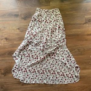 Somrig omlottkjol  - Säljer en vit långkjol från NLY Trend med ett romantiskt blommigt mönster i rött och rosa. Kjolen har en flowy passform och är tillverkad i ett lätt, luftigt material som ger en skön känsla. Perfekt för dig som gillar feminina och somriga vibes.