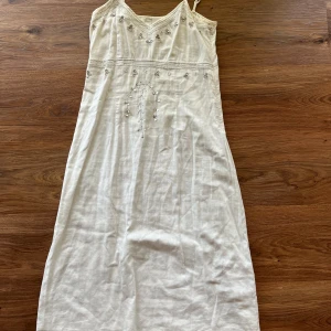 Gullig vit klänning - Vit klänning från Fransa med smala axelband och v-ringning som går ungefär till knäna. Klänningen har broderade blommor och små paljetter som ger en glittrig touch. Tillverkad i ett lätt material, perfekt för varma dagar. Snygg och feminin design med fina detaljer.