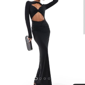 Nyårsklänning Svart långklänning med twist och cut out - Supersnygg svart långklänning med twistad detalj framtill och stor cut out över magen. Klänningen har lång ärm och figurnära passform som framhäver siluetten. Perfekt för dig som vill sticka ut med en modern och edgy look. Helt ny