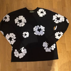 Svart långärmad tröja Denim Tears - Svart långärmad tröja från Denim Tears med stora vita blommiga tryck över hela plagget. Tröjan har en avslappnad passform och är tillverkad i mjuk bomull. Perfekt statement-piece för dig som vill sticka ut med coolt mönster. ⚠️1:1⚠️