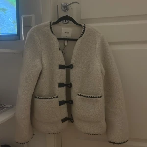 Vit teddyjacka från Object, strl 34 - Supermysig vit teddyjacka från Object med svarta detaljer och dekorativa knappar framtill. Jackan har två fickor med svart kant och coola snörningsdetaljer. Perfekt för dig som vill ha en trendig och varm look i höst och vinter.