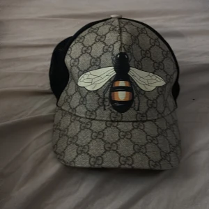 Gucci keps med bi och GG-mönster - Säljer en snygg Gucci keps med klassiskt GG-mönster i beige och grå toner. Framtill finns en stor bi-print i svart, vitt och orange. Baksidan är i svart mesh för extra ventilation. Märkt med Gucci-label och tillverkad i bomull och polyester. Kan gå ner i pris