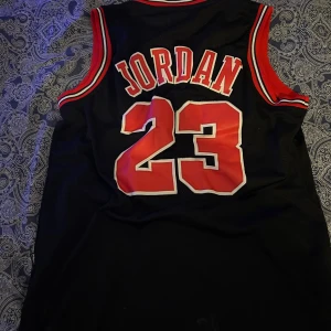 Chicago Bulls Jordan 23 Nike tröja - Säljer en svart Chicago Bulls baskettröja från Nike med Michael Jordans namn och nummer 23 i rött och vitt. Tröjan är ärmlös med röda och vita detaljer vid hals och ärmhål, tillverkad i mesh-material för extra ventilation. Perfekt för dig som älskar basket och streetwear.