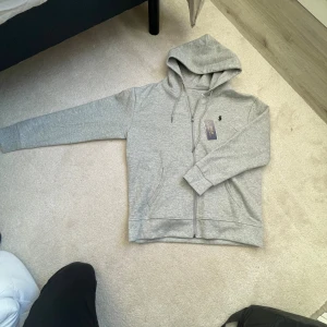 Grå hoodie från Polo Ralph Lauren - Säljer en stilren grå hoodie från Polo Ralph Lauren med dragkedja, huva och snörning. Tröjan har lång ärm, två fickor fram och den klassiska Polo-loggan broderad på bröstet. Perfekt för chill dagar och enkel att matcha med jeans eller joggers.
