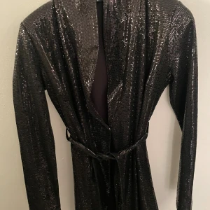 Svart glittrig långklänning med bälte - Svart långklänning med glittrigt tyg och lång ärm. Klänningen har ett bälte i midjan och en öppen front som ger en snygg och trendig look. Perfekt för dig som vill sticka ut med en elegant och cool stil.