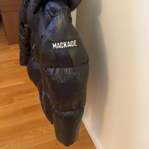 Svart pufferjacka från Mackage - Säljer en svart pufferjacka från Mackage med glansig finish och huva. Jackan har två snedställda fickor med dragkedja och tydlig logga på ärmen. Perfekt för kalla dagar och riktigt snygg streetstil. Passar mig som har storlek s/m