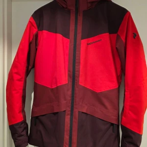 Röd och vinröd vindjacka Peak Performance - Snygg vindjacka från Peak Performance i rött och vinrött med svart logga på bröstet. Jackan har huva, hel dragkedja framtill och praktiska fickor. Perfekt för dig som gillar stilren och sportig look. Finns även byxor till som ej fick plats i bilderna