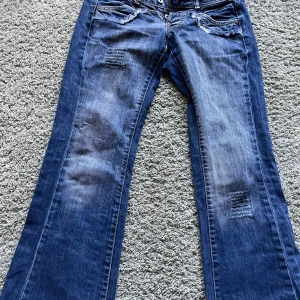 Blå bootcut jeans från Glago - Snygga blå jeans från Glago med bootcut passform och låg midja. Jeansen har slitna detaljer vid fickorna, patchade partier på benen och dubbla knappar i midjan. Materialet är klassisk jeans med en cool tvättad look och fransade kanter.