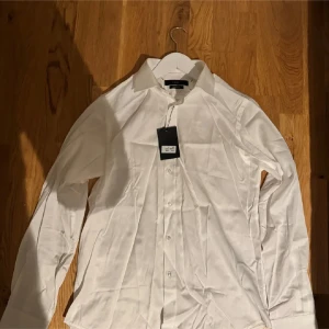 Vit skjorta från POUR Regular Fit M - Klassisk vit skjorta från POUR i regular fit med lång ärm och knappar framtill. Skjortan har en stilren krage och är tillverkad i ett slätt tyg som passar perfekt till en clean och snygg look.