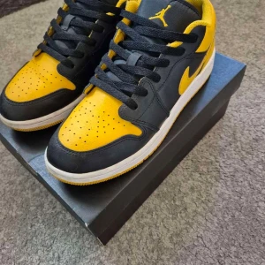 Nike Air Jordan 1 Low svart/gul - Nike Air Jordan 1 Low sneakers i svart och gul med vit sula. Klassisk låg modell med snörning och perforerad tå. Ikonisk Jumpman-logga på plösen och Swoosh på sidan. Skorna är tillverkade i skinn och har en rund tå och platt sula.