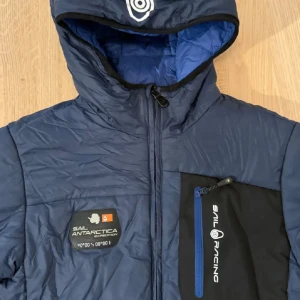 Blå Sail Racing pufferjacka med huva - Snygg blå pufferjacka från Sail Racing med huva och logga på bröstet. Jackan har svarta detaljer, dragkedjor och en bröstficka med Sail Racing-tryck. Perfekt för kyliga dagar och har en sportig look med expedition badge på bröstet. Dunfoder och yttermaterial är vattentätt och andas.