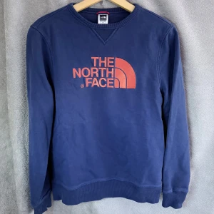 Blå sweatshirt från The North Face - Mörkblå sweatshirt från The North Face med röd logga broderad på bröstet och liten logga på ryggen. Tröjan har rund hals, ribbade muddar och är tillverkad i mjuk bomull. Perfekt för dig som gillar en clean och sportig stil.