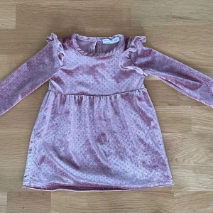 Rosa prickig sammetsklänning Sinsay - Superfin rosa sammetsklänning från Sinsay, Box & Bunny, med prickigt mönster och volangdetaljer vid axlarna. Klänningen har lång ärm och rund hals med liten öppning bak. Perfekt för dig som gillar mjuka material och gulliga detaljer.