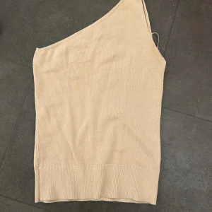 Beige stickad oneshoulder topp - Säljer en beige stickad topp med oneshoulder-design och smala axelband. Toppen har en ribbad kant nedtill och är helt slät utan mönster. Perfekt för dig som gillar stilrena och enkla plagg med en twist.