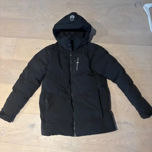 Svart dunjacka från Sail Racing - Svart dunjacka från Sail Racing med huva och dragkedja framtill. Jackan har två sidofickor och en bröstficka med dragkedja, samt logga på bröstet och ärmen. Perfekt för kalla dagar med sin vadderade design och stilrena look.