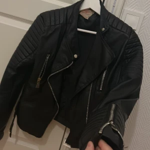 Svart skinnjacka från Voyelles - Snygg svart skinnjacka från Voyelles med coola quiltade detaljer på axlarna, dragkedjor framtill och vid ärmslut samt spännen nedtill. Jackan har en klassisk bikerstil och är perfekt för dig som vill ha en edgy look.